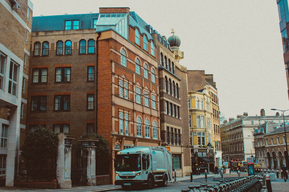A compact street scene featuring a white garbage collection vehicle positioned on the pavement next to a row of multi-story buildings with mixed architectural styles. The vehicle, likely used by independent waste removal services, has a smooth, rounded body with visible side mirrors and black tires, indicating its function for rubbish collection. The buildings behind are composed of various materials, including red brick and painted finishes, with large windows, some arched, and decorative facades, suggesting historical or Victorian influence. The scene includes a partial view of a metal fence or barrier at the roadside, and additional urban infrastructure such as street lamps, bollards, and parked bikes, emphasizing a typical city environment. Natural daylight illuminates the scene, with clear visibility of textures on the vehicle and buildings, reflecting a professional waste management approach for private rubbish clearance on busy urban streets, aligned with services like those offered by rubbishremovalshepherdsbush.org.uk to handle waste efficiently in city environments.