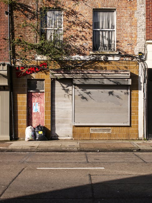 A compact street scene featuring a white garbage collection vehicle positioned on the pavement next to a row of multi-story buildings with mixed architectural styles. The vehicle, likely used by independent waste removal services, has a smooth, rounded body with visible side mirrors and black tires, indicating its function for rubbish collection. The buildings behind are composed of various materials, including red brick and painted finishes, with large windows, some arched, and decorative facades, suggesting historical or Victorian influence. The scene includes a partial view of a metal fence or barrier at the roadside, and additional urban infrastructure such as street lamps, bollards, and parked bikes, emphasizing a typical city environment. Natural daylight illuminates the scene, with clear visibility of textures on the vehicle and buildings, reflecting a professional waste management approach for private rubbish clearance on busy urban streets, aligned with services like those offered by rubbishremovalshepherdsbush.org.uk to handle waste efficiently in city environments.