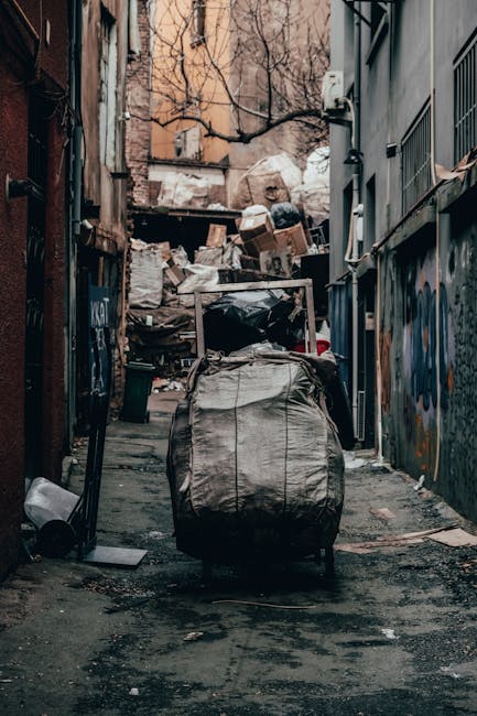 A narrow urban alleyway filled with piles of mixed waste materials, including large black and grey garbage bags, cardboard boxes, and assorted refuse stacked against a weathered wooden fence at the far end. The ground is dark and uneven, with visible patches of dirt and debris. On the left side, part of a building with exposed brickwork and graffiti is visible, while on the right side, a tall grey wall features metal grilles and a small window. In the foreground, a large, wheeled trash bin with a worn fabric cover sits centrally, suggesting an independent rubbish collection or private waste handling method as part of a local rubbish removal service. The scene is lit with natural light, creating a slightly gloomy atmosphere typical of an area prioritizing rubbish clearance services such as those offered by companies like rubbishremovalshepherdsbush.org.uk, emphasizing the need for effective waste management in densely populated urban environments.