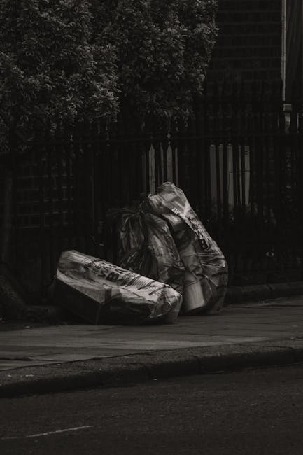 A narrow urban alleyway filled with piles of mixed waste materials, including large black and grey garbage bags, cardboard boxes, and assorted refuse stacked against a weathered wooden fence at the far end. The ground is dark and uneven, with visible patches of dirt and debris. On the left side, part of a building with exposed brickwork and graffiti is visible, while on the right side, a tall grey wall features metal grilles and a small window. In the foreground, a large, wheeled trash bin with a worn fabric cover sits centrally, suggesting an independent rubbish collection or private waste handling method as part of a local rubbish removal service. The scene is lit with natural light, creating a slightly gloomy atmosphere typical of an area prioritizing rubbish clearance services such as those offered by companies like rubbishremovalshepherdsbush.org.uk, emphasizing the need for effective waste management in densely populated urban environments.