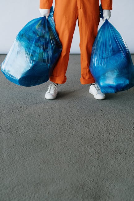 A person wearing orange work overalls and white sneakers is standing on a grey pavement surface, holding two large blue plastic rubbish bags, one in each hand. The bags are translucent, revealing various types of waste materials inside, and are pulled tightly with creases and folds, indicating they are filled with rubbish. The background is plain and grey, suggesting an indoor or outdoor urban environment, possibly a driveway or loading area. The scene depicts a waste collection or rubbish removal activity, aligned with the context of private waste handling or excess waste disposal, which may be managed through independent collection services like those offered by rubbishremovalshepherdsbush.org.uk or similar providers in Shepherds Bush. The focus remains on the manual handling of waste in large, durable plastic bags, highlighting the typical approach for rubbish removal services that operate outside local authority collection methods.